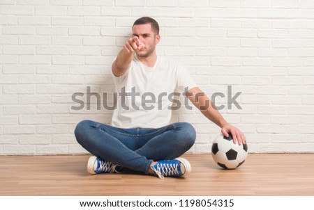 Young caucasian man sitting over white brick wall holding soccer football ball pointing with finger to the camera and to you, hand sign, positive and confident gesture from the front