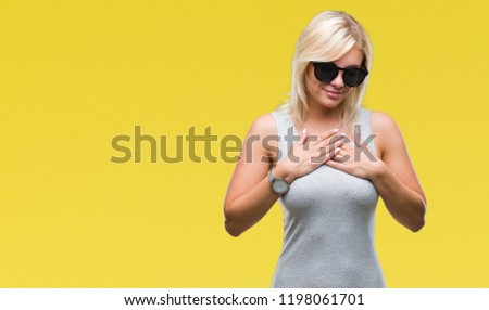 Young beautiful blonde woman wearing sunglasses over isolated background smiling with hands on chest with closed eyes and grateful gesture on face. Health concept.