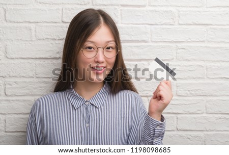 Young Chinese woman over brick wall holding credit card with a happy face standing and smiling with a confident smile showing teeth