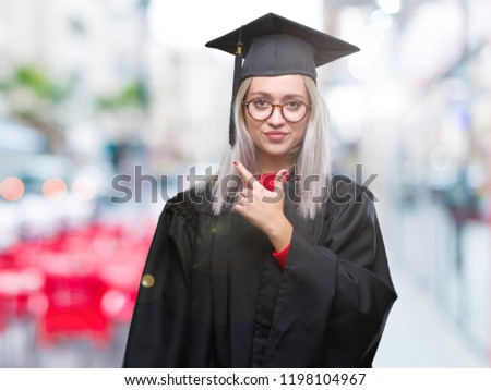 Young blonde woman wearing graduate uniform over isolated background cheerful with a smile of face pointing with hand and finger up to the side with happy and natural expression on face