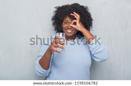 Young african american woman over grey grunge wall drinking a glass of water with happy face smiling doing ok sign with hand on eye looking through fingers