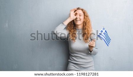 Young redhead woman over grey grunge wall holding flag of Greece with happy face smiling doing ok sign with hand on eye looking through fingers