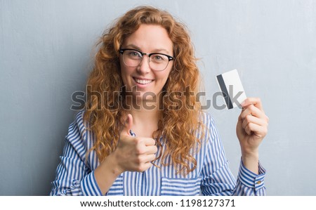 Young redhead woman over grey grunge wall holding credit card happy with big smile doing ok sign, thumb up with fingers, excellent sign