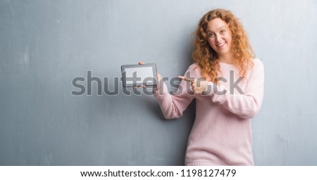 Young redhead woman over grey grunge wall using tablet very happy pointing with hand and finger