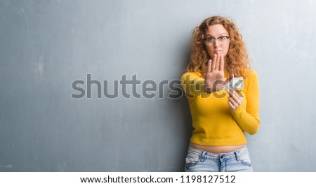 Young redhead woman over grey grunge wall holding a dollar with open hand doing stop sign with serious and confident expression, defense gesture