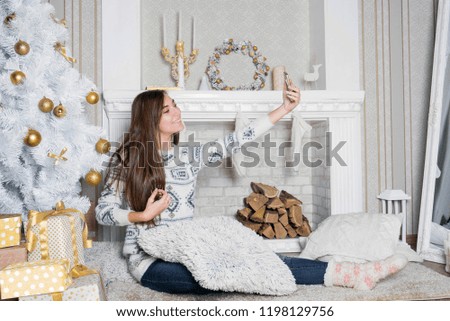 Young attractive brunette woman near the fireplace, cosiness home interior. Teenage girl in white sweater makes selfie. Winter holidays concept. White beautiful decor. Sincere smile. Christmas tree.