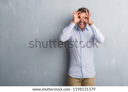 Handsome young business man over grey grunge wall wearing elegant shirt doing ok gesture like binoculars sticking tongue out, eyes looking through fingers. Crazy expression.