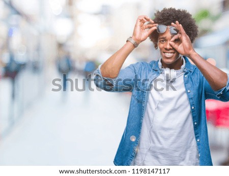 Afro american man wearing headphones listening to music over isolated background doing ok gesture with hand smiling, eye looking through fingers with happy face.