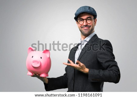 Handsome modern man with beret and glasses holding a piggybank on grey background