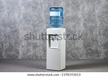White water cooler gallon in office against gray textured wall background. International Exhibition furniture elements in large warehouse interior. Royalty-Free Stock Photo #1198193653