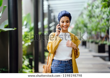 Portrait of a smiling, young and attractive Malay Muslim woman wearing a turban (hijab, head scarf) talking animatedly on her smartphone. She is fashionably dressed and walking in a green city.