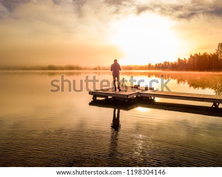 operator of drone drives it to to film foggy bright sunrise over lake and trees, standing with his back to us. Västerbotten,  West Bothnia province, north of Sweden