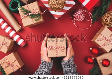 Christmas background with gift boxes, clews of rope, paper's rools and decorations on red. Preparation for holidays. Top view with copy space. Woman's hands holding gift box.