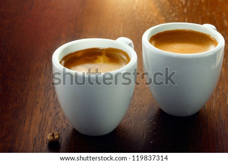 Two espresso coffees in small white cups,with a single coffee bean resting on the wood background