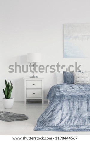 Vertical view of bedroom with big comfortable bed with blue bedding, white bedside table and green plant in pot, real photo with copy space