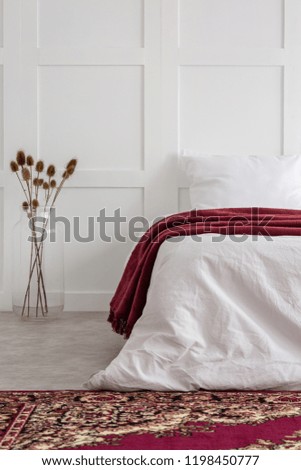 Flowers next to bed with red blanket in white bedroom interior with patterned carpet. Real photo