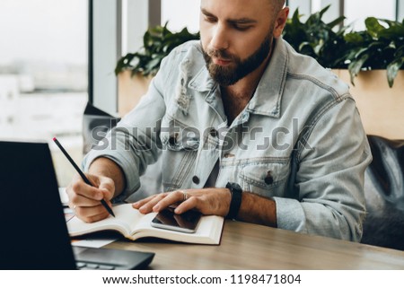 Serious bearded hipster man sitting in office at desk, making notes in notebook, working on laptop.Entrepreneur analyzes information, develops business plan.Freelancer works in cafe. Student learning.