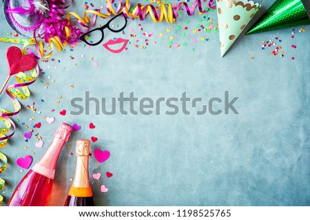 Carnival or New Years border background with colorful streamers, party hats, confetti, accessories and two bottles of champagne on a textured blue grey background with copy space