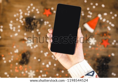 Female hands holding smart mobile phone with oled display on wooden background with Christmas gifts snowflakes and snow magic fairy tale light effect. Happy New Year and Xmas Flat lay Royalty-Free Stock Photo #1198544524