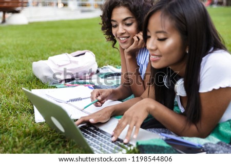 Photo of happy smiling young multiethnic friends girls doing homework using laptop outdoors in park.