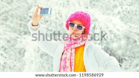 Winter portrait of young woman taking selfie with smartphone  wearing knitted sweater, hat and scarf in the park on snowy trees background