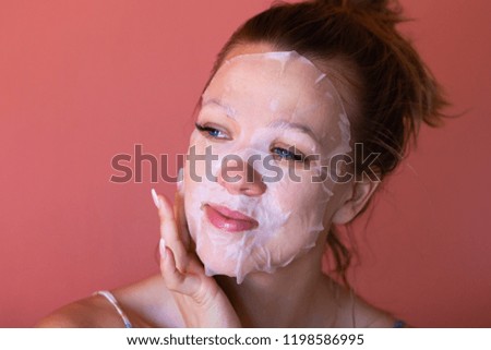 Young woman with blue eyes and white facial mask dreamy smiling and looking at horizon. Pink background. Cosmetic procedure for skin care. Beauty treatments, spa and cosmetology.