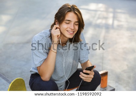 Joyful young guy spending time at the skate park, listening to music with earphones