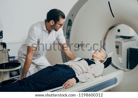 Young medical technician talking to his patient lying on the CT scanner bed.