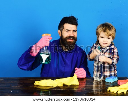 Family time concept. Kid with father holds cleaning sprays. Guy with beard and mustache and kid in checkered shirt. Man with smile and child at brown wooden table on blue background.
