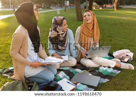 Photo of a happy young arabian women students using laptop computer and holding books in park.