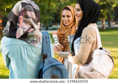 Photo of a cheerful young arabian women students holding books in park outdoors talking with each other.