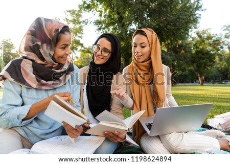 Photo of a happy young arabian women students using laptop computer and holding books in park.