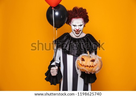 Clown angry man wearing black costume and halloween makeup looking at camera isolated over yellow background holding balloons