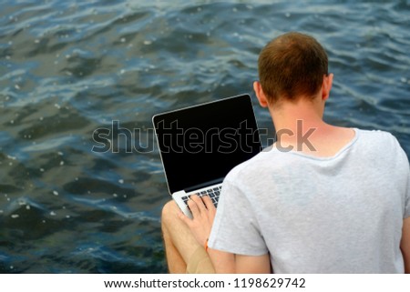 a man with a computer and a telephone in his hands is sitting on the steps near the water, the topic of freelancing and working behind the commuter
