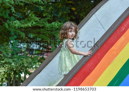 Young girl with golden curly hair, wearing a yellow dress, holds the railing as she climbs a stairway painted with a rainbow on its side