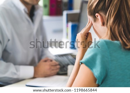 Woman suffering from bad headache or migraine. Appointment with doctor in office room. Sick and unwell lady with stress, trauma or burnout. Young patient in pain holding head with hands. Medical visit Royalty-Free Stock Photo #1198665844