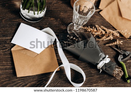 There are little bouquet of flowers, wineglass, postcard, envelopes and white ribbon on the wooden table