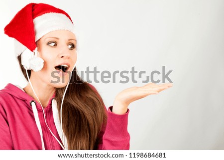 Attractive smiling young girl dressed in Santa's hat listening the music by headphones. Christmas and New Year advertising concept. Detailed studio shoot isolated on abstract blurred white background