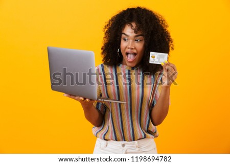 Photo of excited shocked african woman posing isolated over yellow background using laptop computer holding credit card.