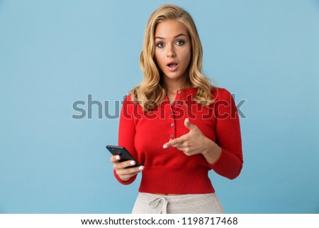 Portrait of european blond woman 20s wearing red shirt pointing at smartphone isolated over blue background in studio