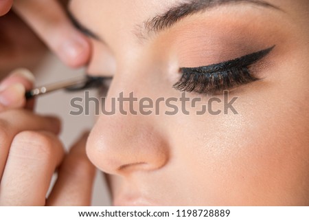 Macro detail of caucasian model false eyelashes during make-up session. The make-up artist is applying a black eyeliner with the brush. The model has white, clean complexion. Royalty-Free Stock Photo #1198728889