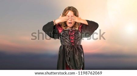 Kid dressed as a vampire at halloween holidays covering eyes by hands at outdoor with sunset