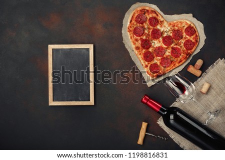 Heart shaped pizza with mozzarella, sausagered with a bottle of wine and wineglas and chalk board. Valentines day greeting card on rusty background . Top view Royalty-Free Stock Photo #1198816831