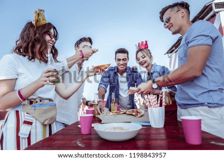 Summer holidays. Happy female person holding bottle in right hand while giving piece of pizza to her friend