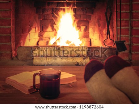 Cup of tea, book, women's feet in warm socks on a wooden table opposite a burning fireplace Royalty-Free Stock Photo #1198872604