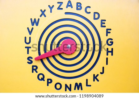 English alphabet in the form of a clock face on a yellow wooden background with a red arrow. Arrow points to the letter "S".