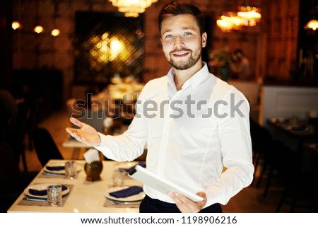 Handsome young friendly man in white shirt holding tablet and greeting with gesture smiling at camera in restaurant