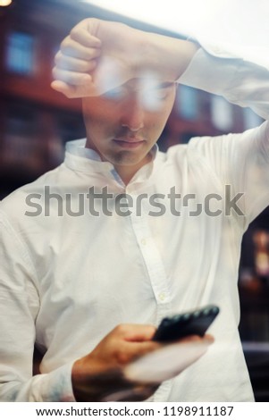 View through shiny glass of modern businessman in white shirt leaning on window and surfing phone