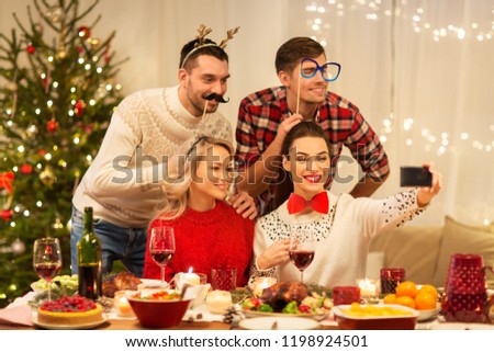 holidays and celebration concept - happy friends with party props taking selfie by smartphone at home christmas dinner