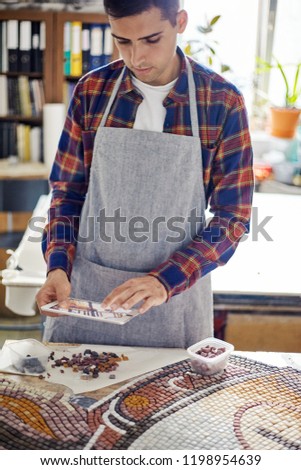 Casual man in apron working with mosaic layout and using tablet to check design 